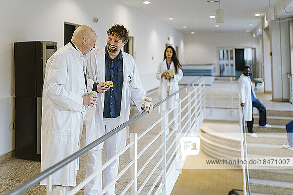 Fröhliche männliche Ärzte  die sich während der Kaffeepause im Krankenhausflur am Geländer stehend unterhalten