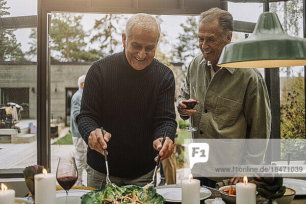 Glücklicher reifer Mann  der mit einem männlichen älteren Freund auf einer Dinnerparty steht und Salat zubereitet