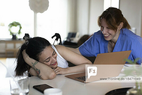 Glückliche Betreuerin im Gespräch mit einer querschnittsgelähmten Frau  die zu Hause auf ihren Laptop schaut