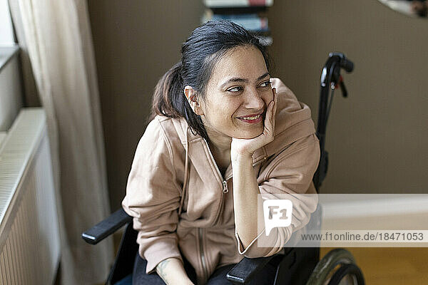 Lächelnde junge Frau mit Hand am Kinn  die zu Hause im Rollstuhl sitzt