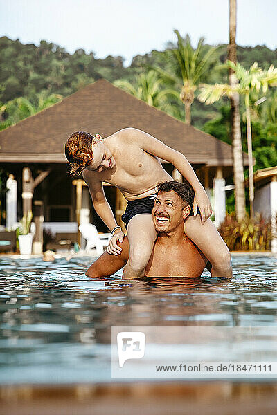 Fröhlicher Vater trägt seinen Sohn auf den Schultern im Schwimmbad eines Ferienortes