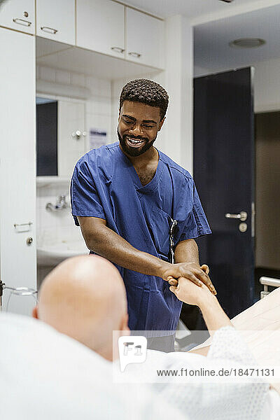 Lächelnder Krankenpfleger hält die Hände eines älteren Patienten  der auf einem Bett im Krankenhaus liegt