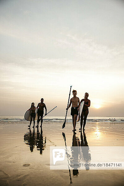 Family with paddleboards walking against sky during sunset at beach