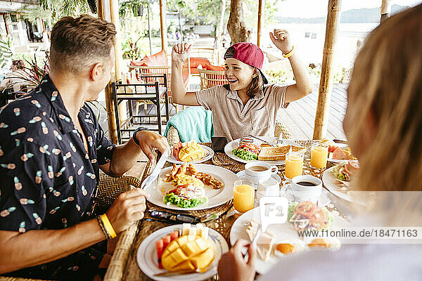 Happy girl clenching fists while parents having breakfast at table in resort