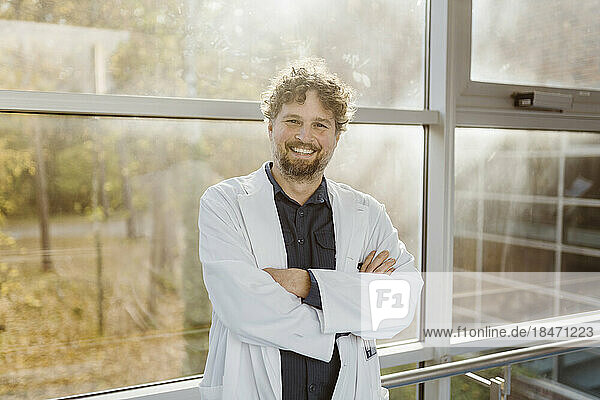 Portrait of smiling mature doctor wearing lab coat standing with arms crossed against window in hospital corridor