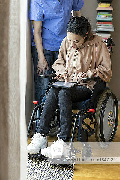 Frau mit Behinderung benutzt Tablet-PC  während sie im Rollstuhl sitzt und von einer Pflegekraft zu Hause betreut wird