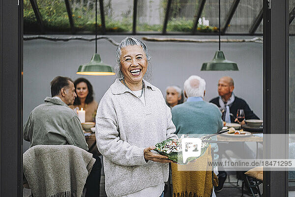 Porträt einer fröhlichen älteren Frau mit einer Salatschüssel in der Hand bei einer Dinnerparty