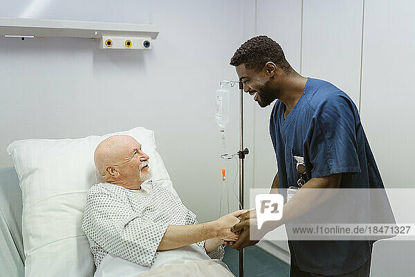 Smiling healthcare worker holding hands of senior male patient lying on bed in hospital