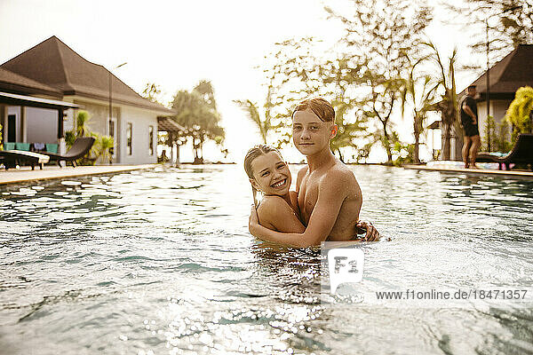 Porträt von Bruder und Schwester  die sich bei Sonnenuntergang im Schwimmbad eines Ferienorts umarmen