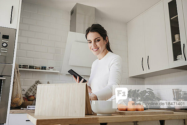 Happy woman using tablet PC holding phone in kitchen