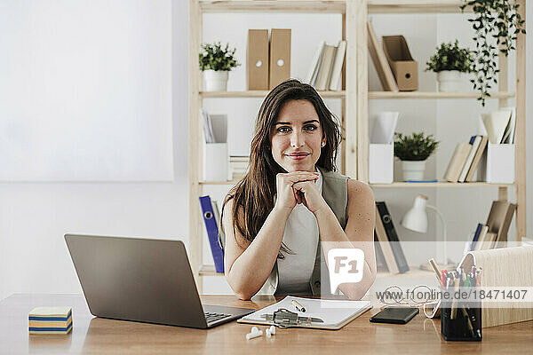 Lächelnde Geschäftsfrau sitzt mit den Händen am Kinn am Schreibtisch im Büro