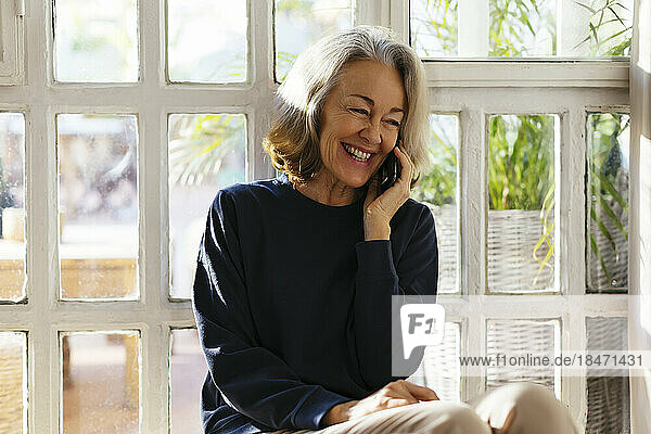 Cheerful mature woman talking on smart phone in front of window