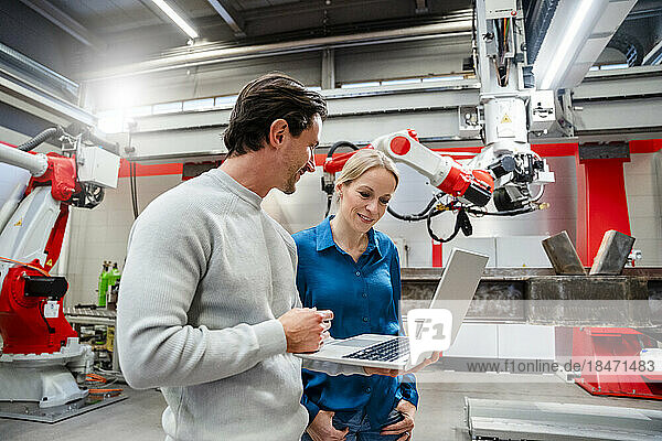 Smiling engineer standing with colleague holding laptop in robot factory