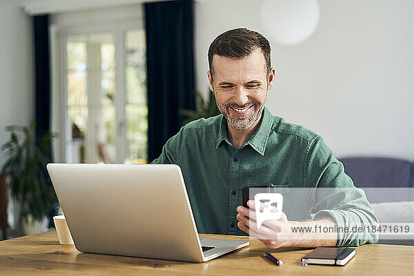 Fröhlicher Mann  der zu Hause am Tisch sitzt und mit dem Smartphone am Laptop bezahlt