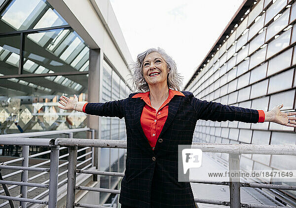 Happy mature businesswoman standing with arms outstretched in front of railing