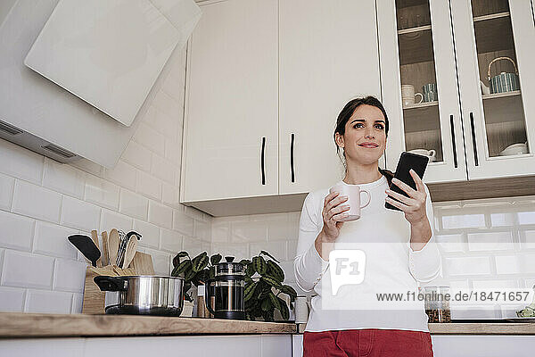 Thoughtful smiling woman with smart phone holding coffee cup in kitchen