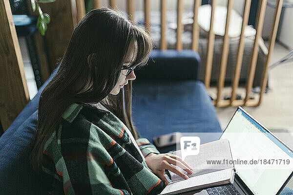 Young woman with laptop reading book at home