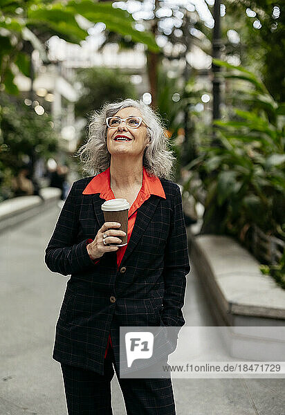 Nachdenkliche reife Geschäftsfrau mit Einwegkaffeetasse steht im Garten