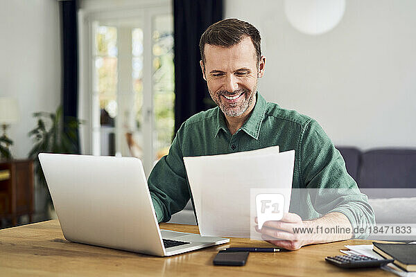 Lächelnder Mann  der von zu Hause aus mit einem Laptop arbeitet und Dokumente in der Hand hält