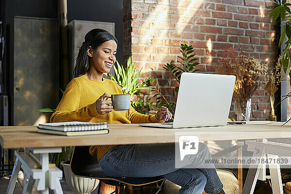 Happy businesswoman with cup doing video call on laptop at loft office