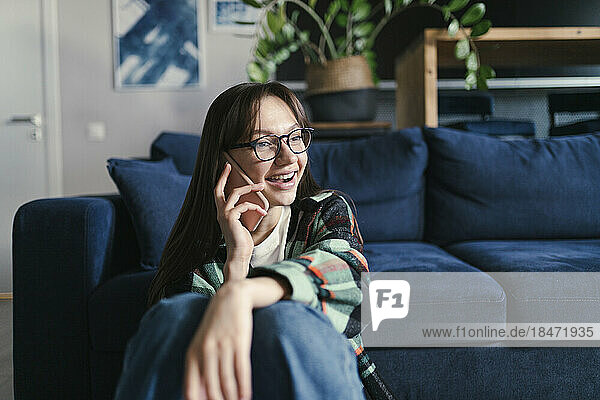 Happy woman talking on smart phone sitting in front of sofa at home