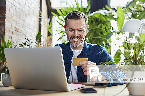 Smiling businessman with laptop holding credit card at loft office