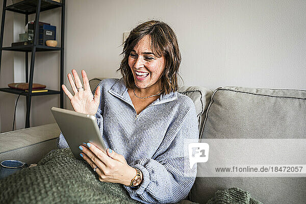 Glückliche Frau winkt per Videoanruf durch den Tablet-PC auf dem heimischen Sofa