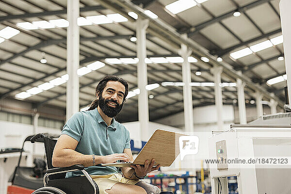 Happy engineer sitting in wheelchair working on laptop in robotics factory