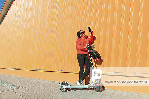 Woman taking selfie through smart phone with electric push scooter standing by wall