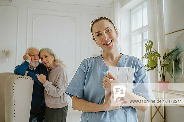 Lächelnde Krankenschwester hält Tablet-PC mit älterem Mann und älterer Frau im Hintergrund zu Hause
