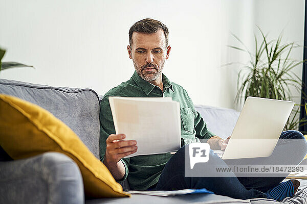 Worried man working from home looking at documents sitting with laptop on sofa