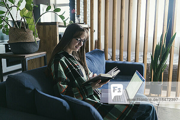 Smiling woman reading book with laptop at home