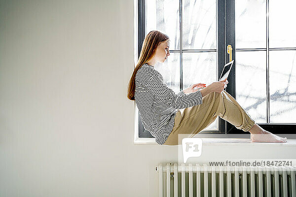 Teenage girl using laptop sitting on window sill at home