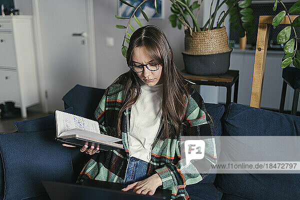 Young woman holding book using laptop sitting on sofa at home