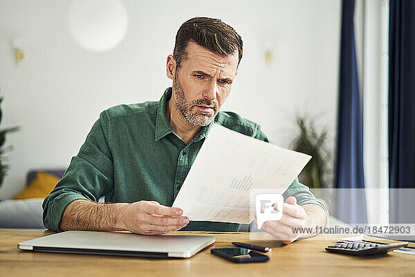 Verärgerter Mann schaut sich Dokument mit Finanzzahlen an  während er zu Hause am Tisch sitzt