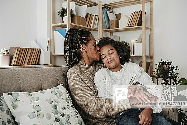 Mother kissing daughter sitting on sofa at home