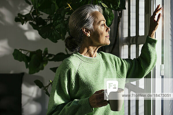 Thoughtful woman with coffee cup standing by window at home
