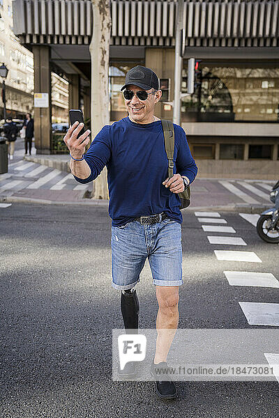 Smiling man with artificial limb taking selfie and crossing road