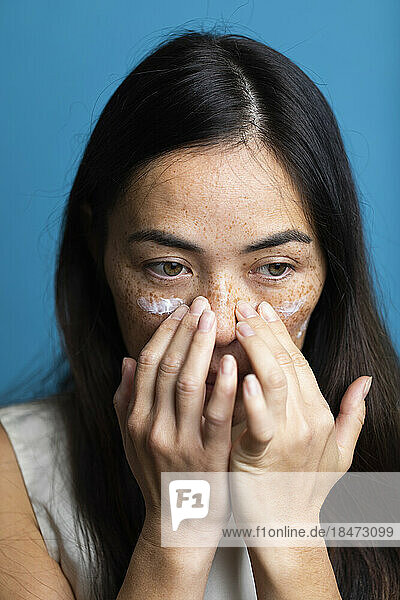 Woman with freckles applying lotion on face against blue background