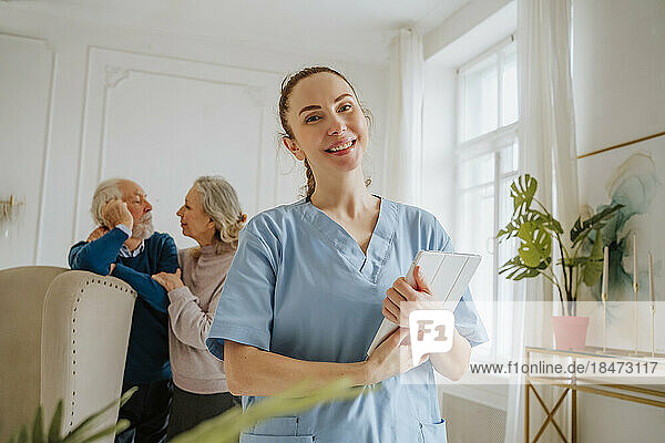 Glückliche Krankenschwester hält Tablet-PC mit älteren Patienten im Hintergrund zu Hause
