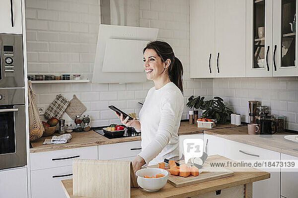 Glückliche Frau steht zu Hause mit Smartphone in der Küche