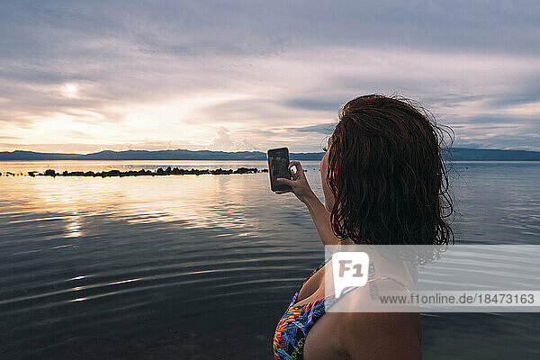 Young woman photographing sunset through smart phone at beach