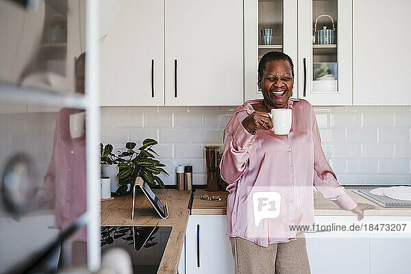 Happy woman holding cup standing in kitchen at home