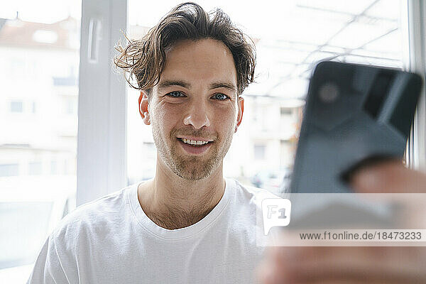 Smiling young man with mobile phone in front of window
