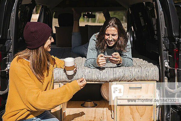 Woman talking to smiling friend using smart phone lying in campervan