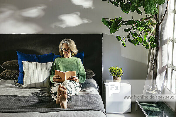 Mature woman reading book sitting on bed at home