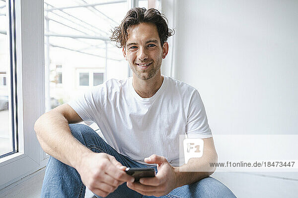 Happy young man sitting with smart phone on window sill