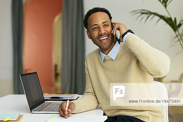 Happy young businessman talking on smart phone at desk in office