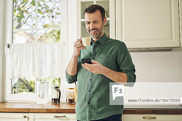 Smiling man using phone while drinking coffee in his kitchen