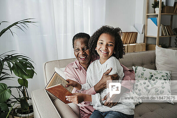 Glückliches Mädchen hält ein Buch in der Hand und sitzt mit Großmutter zu Hause auf dem Sofa